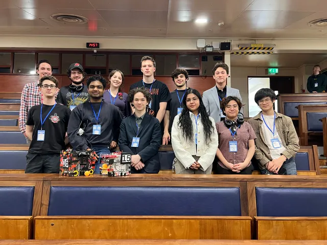 Thirteen society members lined up in a lecture hall at Unibots UK 2026, two competition robots on the bench in front.