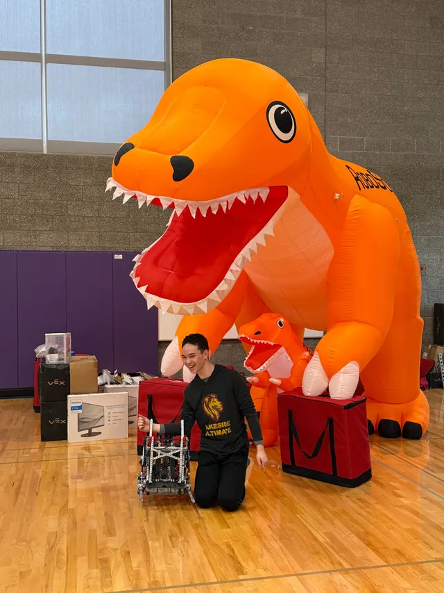 Kellan kneeling next to the state-championship robot, with a giant inflatable dinosaur competition mascot.